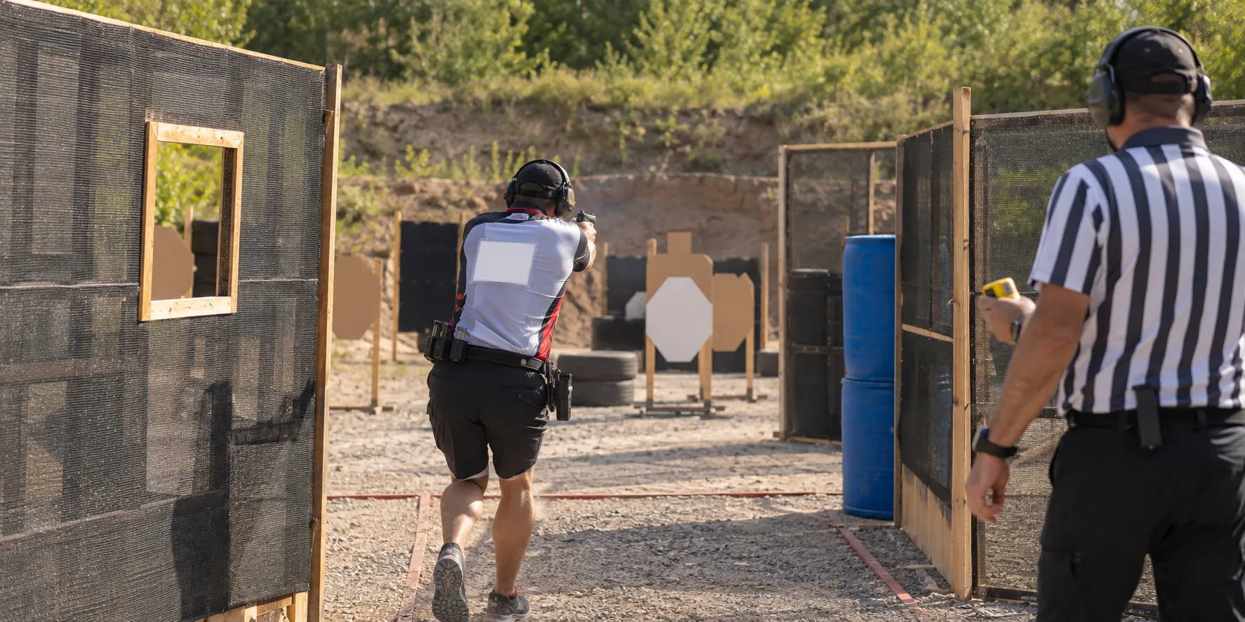 Erwachsener Teilnehmer und Aufsicht bei einem IPSC-Wettkampf auf einem Schießstand, von hinten gesehen
